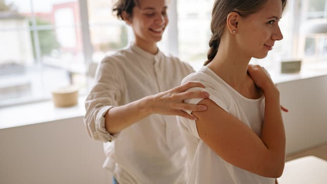 A massage therapist with her hand on a smiling client's shoulder, checking how much pressure feels right.
