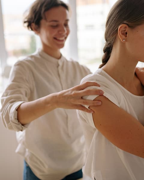 A massage therapist with her hand on a smiling client's shoulder, checking how much pressure feels right.