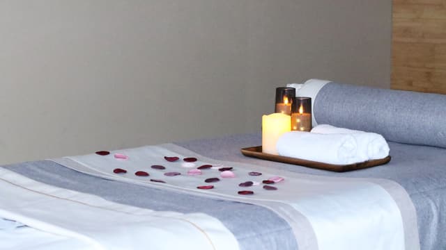 A massage table made up at Wonder Spa with grey and white linens, a bolster, a rolled towel, a candle tray and rose petals.