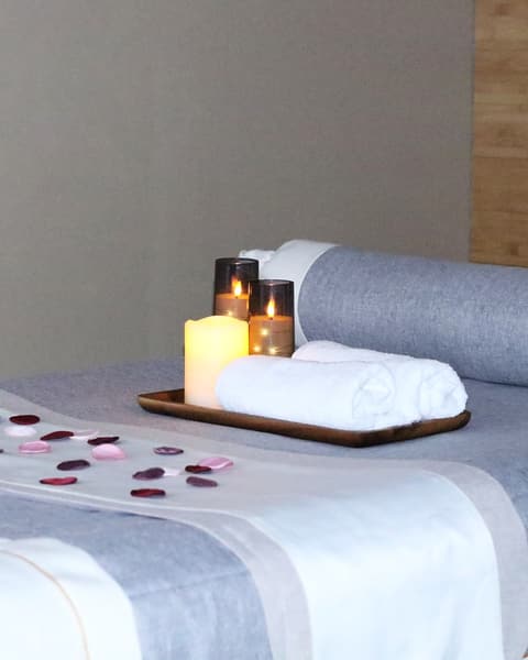 A massage table made up at Wonder Spa with grey and white linens, a bolster, a rolled towel, a candle tray and rose petals.